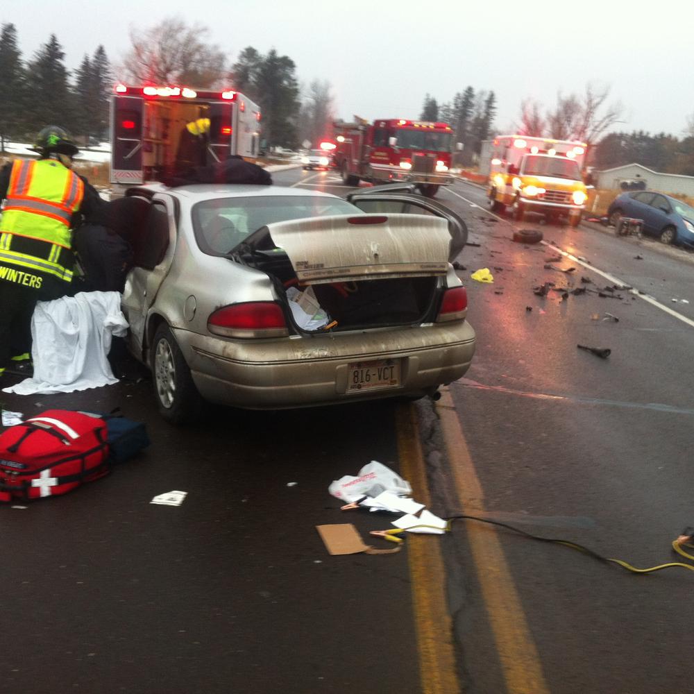 emergency vehicles in the background with crash remnants on the road, vehicle in foreground with trunk and doors open