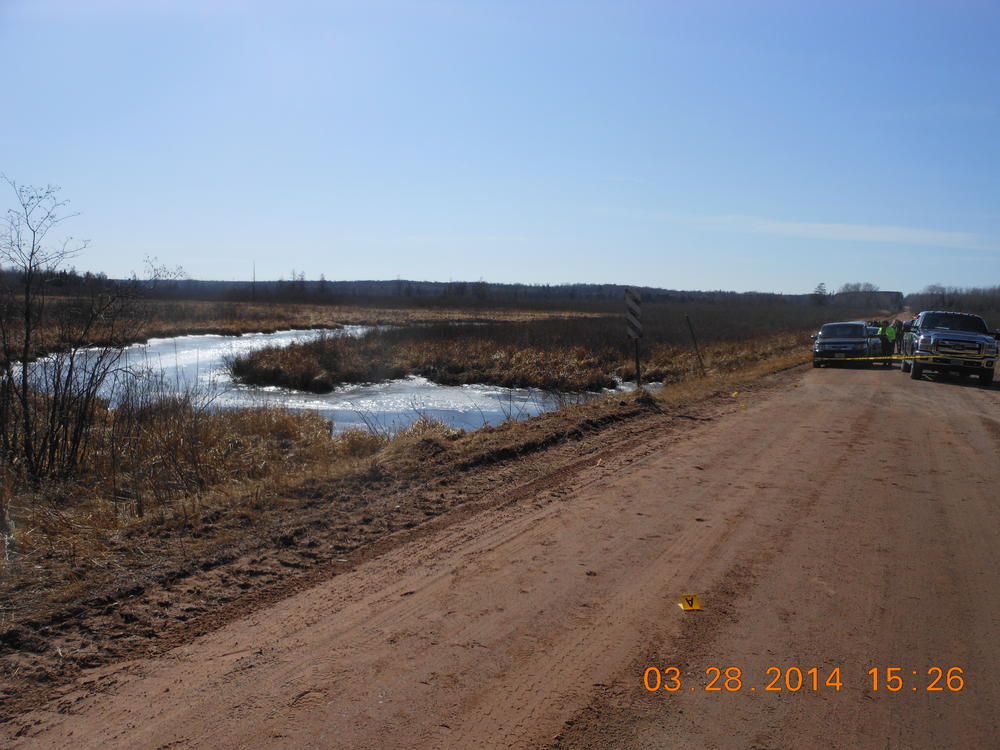 Dirt road with vehicles on the opposite side of a police DO NOT CROSS line, water rushing under a dirt bridge